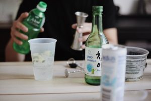 A person is pouring a drink while bottles of Soju and cups sit on the table